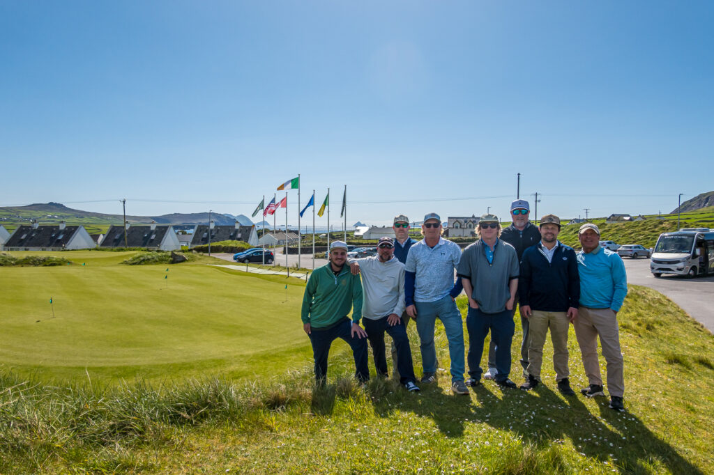 Seven golfers gather beside a putting green and international flags at an Irish clubhouse