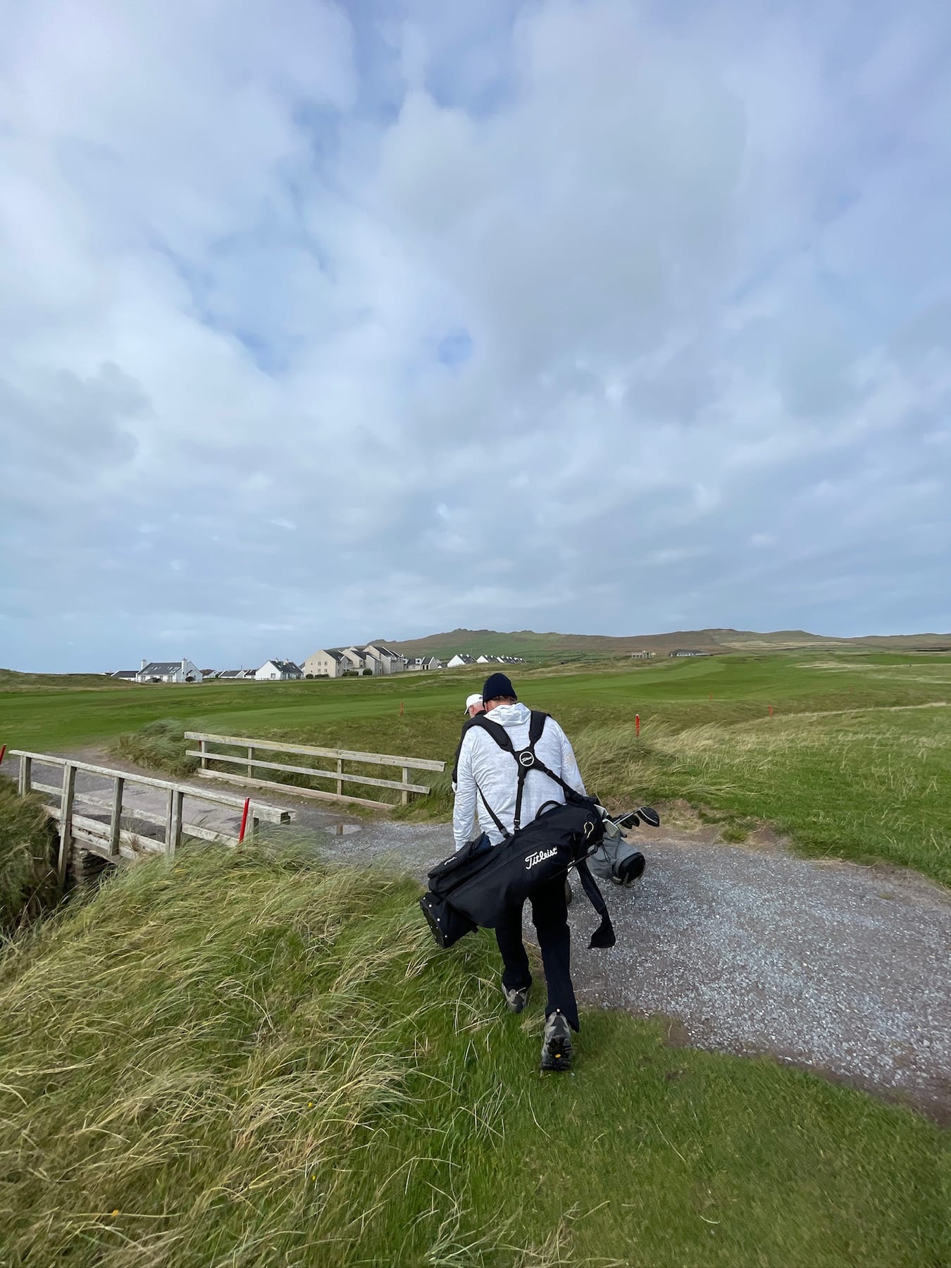 A golfer carrying a bag along a course path beneath a cloudy Irish sky